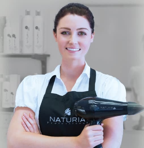 Hair stylist holding a hair dryer, wearing a Naturia apron, and Naturia products in the background on a shelf